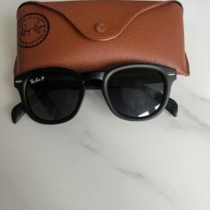 Svarta solglasögon från Ray-Ban - Snygga svarta solglasögon från Ray-Ban med runda, breda bågar och mörka polariserade glas. Kommer med ett klassiskt brunt Ray-Ban-fodral. Perfekt accessoar för soliga dagar.