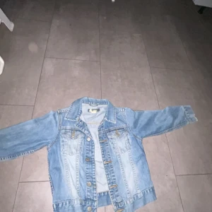 Ljusblå jeansjacka från Kappahl - Säljer en klassisk ljusblå jeansjacka från Kappahl i storlek 134. Jackan har två bröstfickor med knappar och snygga sömdetaljer. Perfekt att slänga över en t-shirt för en cool look.