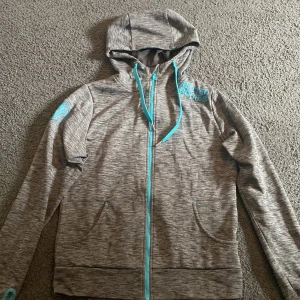 Grå hoodie från Better Bodies - Säljer en gråmelerad hoodie från Better Bodies med turkosa detaljer. Träningshoddien har huva, lång ärm och tryckt logga på bröstet och ärmen. Luftigt och bra material, varan är i bra skick.