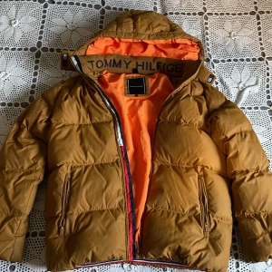 Brun dunjacka från Tommy Hilfiger - Säljer en brun pufferjacka från Tommy Hilfiger med orange foder och tydlig logga på insidan av kragen. Jackan har dragkedja och två fickor framtill. Perfekt för kyliga dagar när du vill hålla dig varm och snygg.