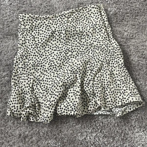 Vit och svart blommig kjol från Zara - Supersöt vit kjol från Zara med små svarta blommor och volang nertill. Kjolen har dragkedja i sida. Lätt och luftig modell som passar till många tillfällen. Inbyggda shorts