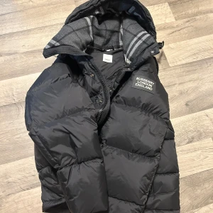 Svart pufferjacka från Burberry - Säljer en svart pufferjacka från Burberry med huva och rutigt foder. Jackan har dragkedja och tryckknappar framtill samt Burberry-logga på bröstet. Perfekt för kyliga dagar och riktigt snygg streetstil.