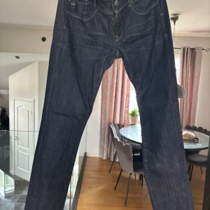 Mörkblå jeansbyxor från G star - Säljer ett par mörkblå jeansbyxor från G star med klassisk femficksdesign och kontrastsömmar. Jeansen har raka ben och normal passform, perfekta till vardags. Snygga detaljer på bakfickorna och knappgylf framtill.