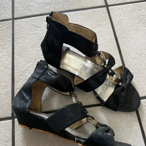 Svarta sandaler med kilklack och gulddetaljer - Säljer ett par svarta sandaler med kilklack och öppen tå. Skorna har dekorativa guldfärgade ringar och små rosetter framtill. Dragkedja baktill för enkel påtagning. Perfekta för dig som vill ha en snygg och bekväm sko med lite extra detaljer.