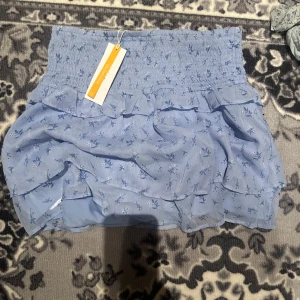 Ljusblå volangkjol med blommönster - Supersöt ljusblå kjol med små blommor och volanger. Kjolen har bred resår i midjan och är luftig och lätt, perfekt för varmare dagar. Den har ett romantiskt och lekfullt uttryck med flera lager volanger.