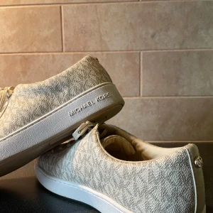 Beige sneakers från Michael Kors - Snygga beige sneakers från Michael Kors med diskret logomönster och vita sulor. Skorna har snörning och guldfärgade detaljer på snörena. Perfekta för en stilren och trendig look.