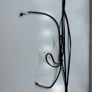 Svart halsband med knyt och silvriga detaljer - Säljer ett svart halsband med tunna band och knyt, dekorerat med silvriga metalldetaljer i ändarna. Perfekt för att ge en stilren touch till din outfit.