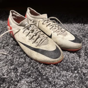 Vit och röda fotbollsskor från Nike - Säljer ett par beige fotbollsskor från Nike med svarta detaljer och röda inslag på sulan. Skorna har snörning och är designade för spel på gräs med fasta dobbar. Perfekta för dig som vill ha stilrena och funktionella skor på planen.