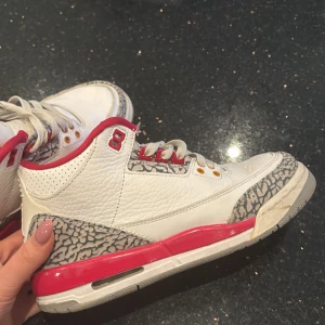 Nike Air Jordans 3 Retro Fire Red - Säljer ett par Nike Air Jordans 3 Retro Fire Red med vit ovandel i skinn, röda detaljer och klassiskt elefantmönster vid tå och häl. Skorna har snörning, rund tå och platt sula. Perfekta för dig som gillar streetwear och sneakers med ikonisk design.