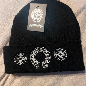 Svart mössa från Chrome Hearts - Svart stickad mössa från Chrome Hearts med vit broderad logga och text både fram och bak. Mössan har uppvikt kant och detaljerade broderier som ger en cool och stilren look.