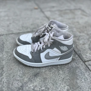 Nike Air Jordan 1 mid wolf grey - Säljer ett par Nike Air Jordans i snygg grå och vit färgkombination. Skorna har klassisk snörning, rund tå och är tillverkade i syntetmaterial med detaljer i skinn. Perfekta för dig som gillar streetwear och vill ha en ikonisk sneaker i garderoben.