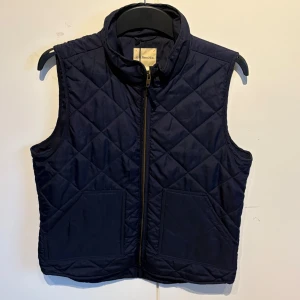 Mörkblå quiltad väst från G.H. Bass & Co. - Stilren mörkblå quiltad väst från G.H. Bass & Co. med dragkedja framtill och två fickor. Västen har ståkrage och är tillverkad helt i polyester, perfekt för lager-på-lager under kyligare dagar. Perfekt till sommaren och våren.