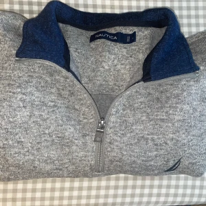 Grå halvzip tröja från Nautica - Säljer en grå långärmad tröja från Nautica med halv dragkedja och mörkblå krage. Tröjan har en broderad logga vid nederkanten och är tillverkad i mjukt material, perfekt för kyligare dagar.