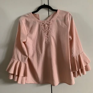 Ljusrosa blus med volangärm från Lindex - Säljer en ljusrosa blus från Lindex med snygga volangärmar och snörning framtill. Blusen har långa ärmar och knäppning i nacken. Perfekt för dig som gillar romantiska detaljer och en feminin stil. stor i storlek så 34 36 fungerar