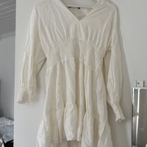 Vit långärmad klänning med v-ringning - Säljer en luftig vit klänning med v-ringad hals och långa ärmar. Klänningen har smockdetaljer över bröstet och volanger nertill för en avslappnad och bohemisk look. Perfekt för varma dagar eller sommarens fester.