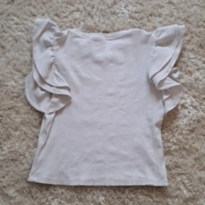 Vit volangtopp från Gina Tricot - Säljer en söt vit topp från Gina Tricot med volangdetaljer på ärmarna. Toppen är ribbad och har en rund halsringning. Perfekt för dig som gillar en feminin och stilren look.