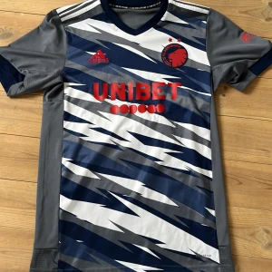 FC Köpenhamn fotbollströja  - Säljer en FC Köpenhamn bortatröja från Adidas med blått och vitt mönster, röda detaljer och sponsortryck. Tröjan har korta ärmar och V-ringning. Perfekt för dig som vill sticka ut på läktaren eller på planen. Den är i storlek S och passar någon mellan 173-183 beroende på vikt! Skriv gärna om ni har några funderingar 😁😁😁