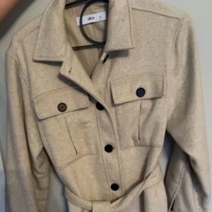 Beige kappa från Ellos - Säljer en beige kappa från Ellos med klassisk krage, knappar framtill och bälte i midjan. Jackan har två bröstfickor med lock och knappar för en snygg detalj. Perfekt för dig som gillar stilrena plagg.