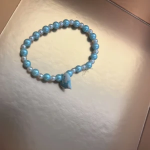Blått pärlarmband med hjärtdetalj - Säljer ett sött armband med ljusblå pärlor och en liten hjärtformad detalj. Perfekt accessoar för att ge lite färg till din outfit.