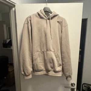 Beige hoodie med ficka - Säljer en beige hoodie med huva och dragsko. Tröjan har en stor magficka och ribbade muddar vid ärmslut och nederkant. Perfekt för en avslappnad stil.