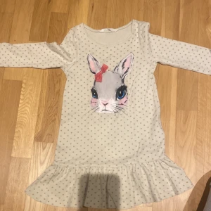 Prickig klänning med kaninmotiv från H&M - Supersöt ljus klänning från H&M med små grå prickar och ett stort kanintryck framtill. Klänningen har volang nertill och långa ärmar. Perfekt för dig som gillar gulliga detaljer och mjuka material.