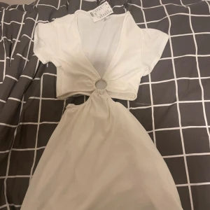 Vit kortklänning med ringdetalj - Säljer en vit kortklänning med djup v-ringning och snygg ringdetalj framtill. Klänningen har korta ärmar och är tillverkad i ett mjukt, lätt material. Perfekt för varma dagar och somriga tillfällen. Perfekt för studenten nu, köpte den för 200
