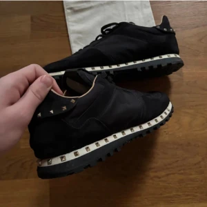 Svarta Valentino Rockrunner sneakers med nitar - Säljer ett par svarta Valentino Rockrunner sneakers med ikoniska silverfärgade nitar runt sulan. Skorna har en kombination av mocka och textil, svart yttersula och rund tå. Perfekta för dig som vill ha en lyxig och edgy look. Inge box. Finns i sthlm