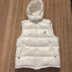 Vit dunväst från Moncler med huva - Säljer en snygg vit dunväst från Moncler med avtagbar huva och två dragkedjefickor. Västen har en klassisk Moncler-logga på bröstet och stängs med dragkedja framtill. Perfekt för lager-på-lager och stilren look.