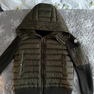 Grön quiltad jacka med huva från Moncler - Snygg grön quiltad jacka från Moncler med huva och dragkedja. Jackan har ribbade muddar, stickade ärmar och en praktisk ficka med dragkedja på ärmen. Perfekt för kyliga dagar och har en modern look.