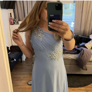 Ljusblå långklänning med paljettdetaljer - Säljer en ljusblå långklänning med oneshoulder och glittriga paljettdetaljer vid axel och midja. Klänningen har draperad framsida och elegant passform, perfekt för bal eller fest. Snygg och unik design som verkligen sticker ut bland gruppen💞💞