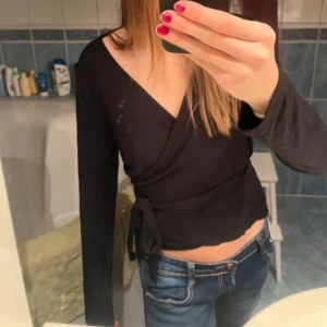 Svart omlottopp med knytband - Säljer en svart omlottopp med långa ärmar och knytband i midjan. Toppen har en djup v-ringning och sitter tight. Perfekt till jeans för en stilren look.
