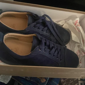Mörkblå sneakers från Christian Louboutin - Säljer ett par mörkblå sneakers från Christian Louboutin med klassisk röd sula. Skorna har snörning och är tillverkade i mocka med svarta detaljer och rund tå. Perfekta för dig som vill ha något stilrent men ändå sticker ut.