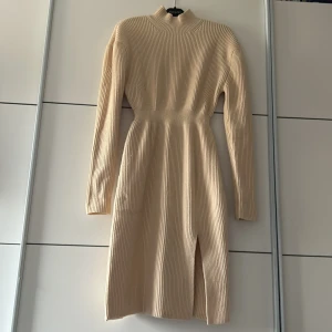 Beige ribbstickad klänning från Adoore - Säljer en beige ribbstickad klänning från Adoore i storlek 40. Klänningen har lång ärm, hög hals och en snygg slits framtill. Den är figurnära och passar perfekt till kyligare dagar. Lite liten i Storleken så passar 38 