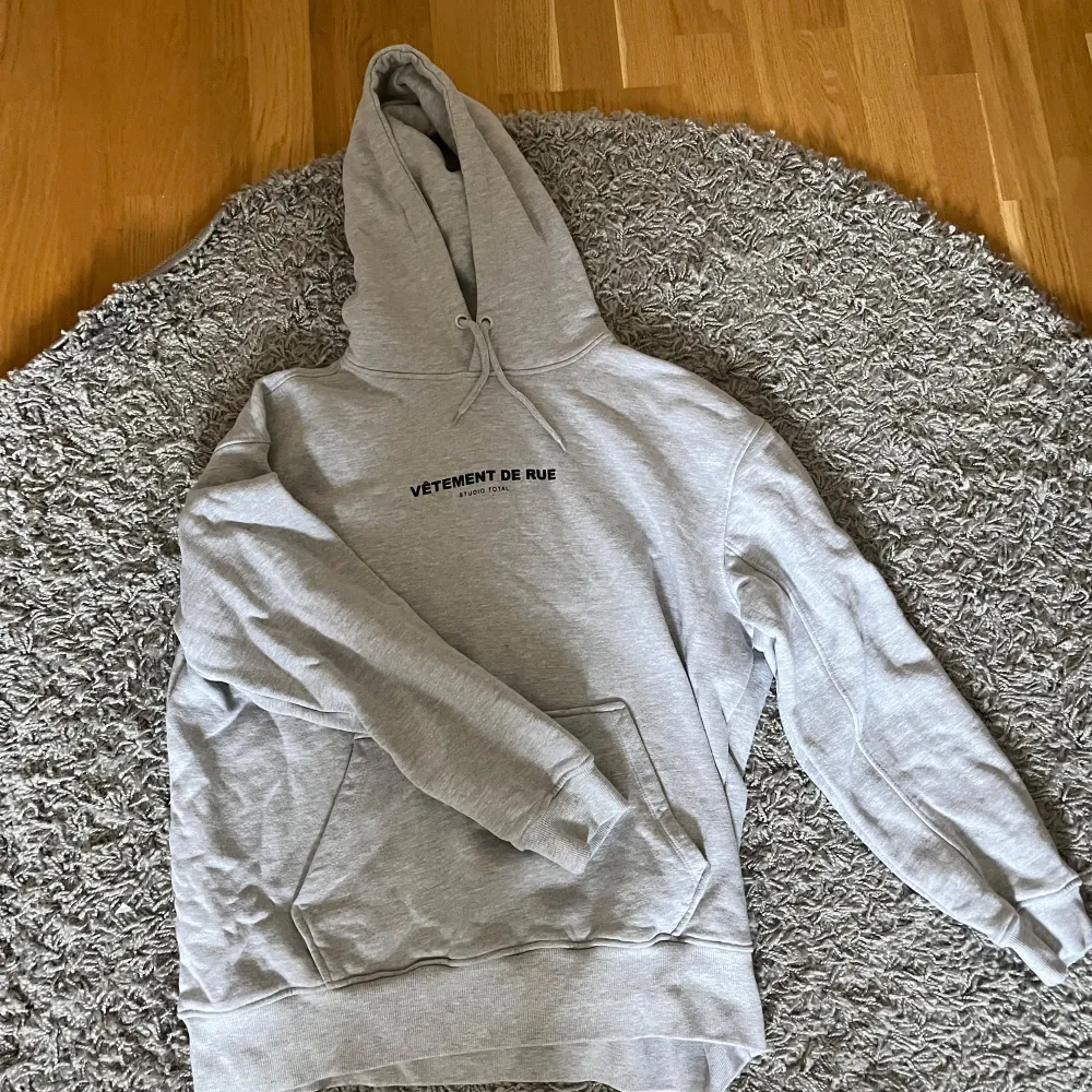 Säljer en ljusgrå hoodie från Studio Total med trycket 'VÊTEMENT DE RUE' på bröstet. Tröjan har huva med dragsko och en stor magficka. Perfekt för en avslappnad stil.. Neuletakit & Villapaidat.
