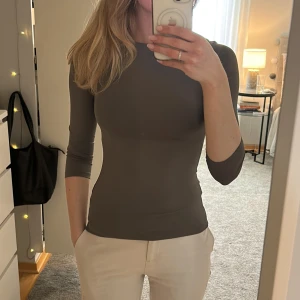 Mörkgrå tight topp med trekvartsärm - Intressekoll! Säljer en mörkgrå, tight topp med trekvartsärm och rund halsringning. Toppen är figurnära och har en stilren, minimalistisk design utan synliga detaljer. Perfekt för dig som gillar en enkel och clean look.