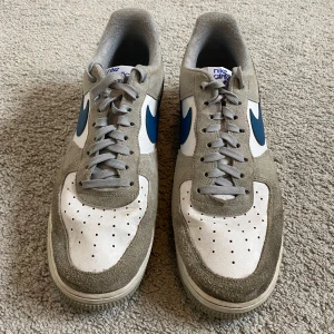 Nike Air Force 1 Athletic Club  - Nike Air Force 1 Athletic Club sneakers i grå mocka och vitt tyg med blå swoosh. Klassisk låg modell med rund tå, perforerad tåbox och platt sula. Grå skosnören och blå detaljer på hälen. Uk size 10 storlek 45. Använd några enstaka gånger.