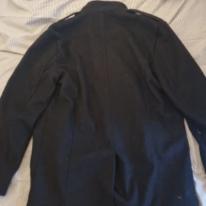 Svart rock med knappar och dragkedja - Säljer en svart rock med klassisk design. Jackan har långa ärmar, knappar framtill och en dragkedja vid bröstet. Perfekt för kyligare dagar och ger en stilren look.