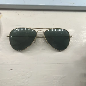 Ray-Ban aviator solglasögon med guldfärgad metallbåge - Klassiska Ray-Ban aviator solglasögon med tunn guldfärgad metallbåge och mörkgröna glas. Snygg och tidlös modell som passar till många stilar. Ray-Ban-loggan syns på linsen. De är väldigt små och rekommenderar därför att de köps till ett barn eller någon med litet ansikte/huvud.