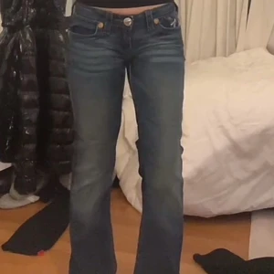 Blå bootcut jeans från True Religion - Säljer ett par klassiska blå jeans från True Religion med bootcut-passform. Jeansen har tydliga kontrastsömmar och ikoniska bakfickor med broderad logga. Märkespatch bak i midjan. Perfekta för dig som gillar en tidlös jeanslook.