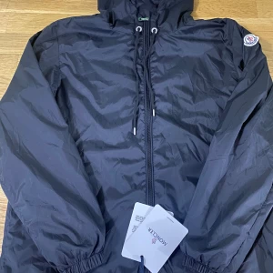 Svart vindjacka från Moncler - Säljer en svart vindjacka från Moncler med huva och dragsko. Jackan har dragkedja framtill, elastiska muddar och Moncler-logga på ärmen. Perfekt för blåsiga dagar och enkel att matcha med det mesta. Storlek S {170,180}