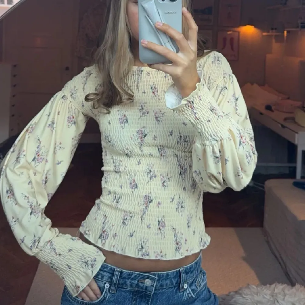 Söt gul blus med blommigt mönster och långa puffärmar. Toppen är tight med smockad midja och ärmslut, samt har en lätt volangkant nertill. Perfekt för en somrig look! Slutsåld överallt!. Yläosat.