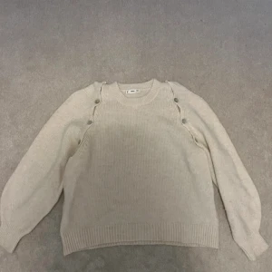 Beige stickad tröja med knappar från MNG - Säljer en beige stickad tröja från MNG med rund halsringning och dekorativa knappar längs ena axeln. Tröjan har långa ärmar och ribbade muddar vid ärmslut och nederkant. Perfekt för lager-på-lager-stil!