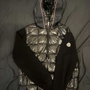 Svart pufferjacka från Moncler med huva - Säljer en svart glansig pufferjacka från Moncler med stickade svarta ärmar och huva. Jackan har dragkedja framtill och Moncler-logga på ärmen. Perfekt för kyliga dagar och snygg streetstil.