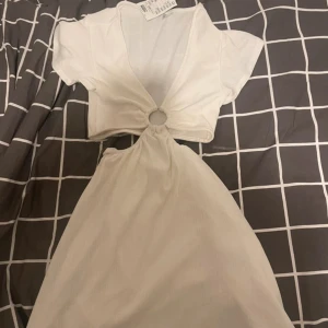 Vit kortklänning med ringdetalj - Superfin vit kortklänning från Bershka med djup v-ringning och snygg ringdetalj framtill. Klänningen har korta ärmar och är gjord i ett lätt ribbat material. Perfekt för varma dagar och en trendig look. Med prislapp, köpte för 200