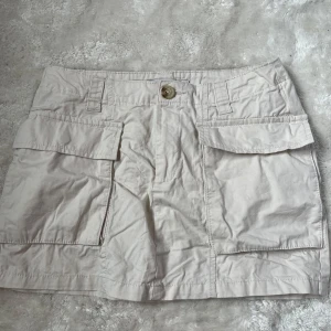 Beige kortkjol med fickor från 157 - Säljer en beige kortkjol från 157 med stora fickor framtill och knappstängning. Kjolen har hällor för skärp och är tillverkad i ett lätt bomullsmaterial. Perfekt för en avslappnad och stilren look. Finns en liten liten fläck vid knappen.