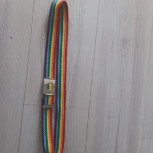 Regnbågsfärgat bälte med hjärtspänne - Säljer ett färgglatt bälte med regnbågsränder och ett unikt hjärtformat spänne i silver. Perfekt för att lägga till en pop av färg till din outfit.
