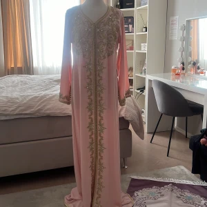 Rosa marockansk kaftan med broderier - Elegant marockansk rosa kaftan långklänning med vackra guldfärgade broderier längs framsidan och ärmsluten. Klänningen har en v-ringad design och långa ärmar, perfekt för speciella tillfällen. Köpt från Spanien och har aldrig kommit till användning.