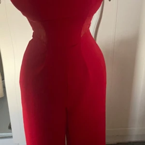 Röd jumpsuit med spetsdetaljer - Säljer en snygg röd långklänning med smala axelband och insydda spetsdetaljer i midjan. Klänningen har en figurnära passform upptill och faller rakt ned. Perfekt för bal eller festliga tillfällen.