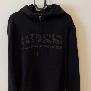 Svart hoodie från BOSS - Säljer en svart hoodie från BOSS med glittrig logga framtill. Tröjan har huva med dragsko och en stor magficka. Perfekt för en chill och stilren look.