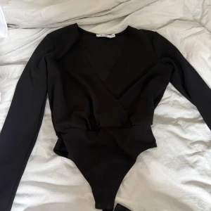 Svart omlott bodysuit från NA-KD - Säljer en svart långärmad bodysuit från NA-KD i omlottmodell. Bodyn har v-ringning och knäppning nedtill. Den har inga defekter förutom på lappen på insidan som syns på bilden 